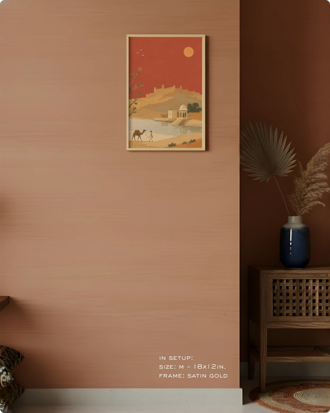 A framed art print featuring a desert landscape with a camel, camel caravan, and a red-orange sky, displayed on a textured terracotta wall in a styled interior setting.