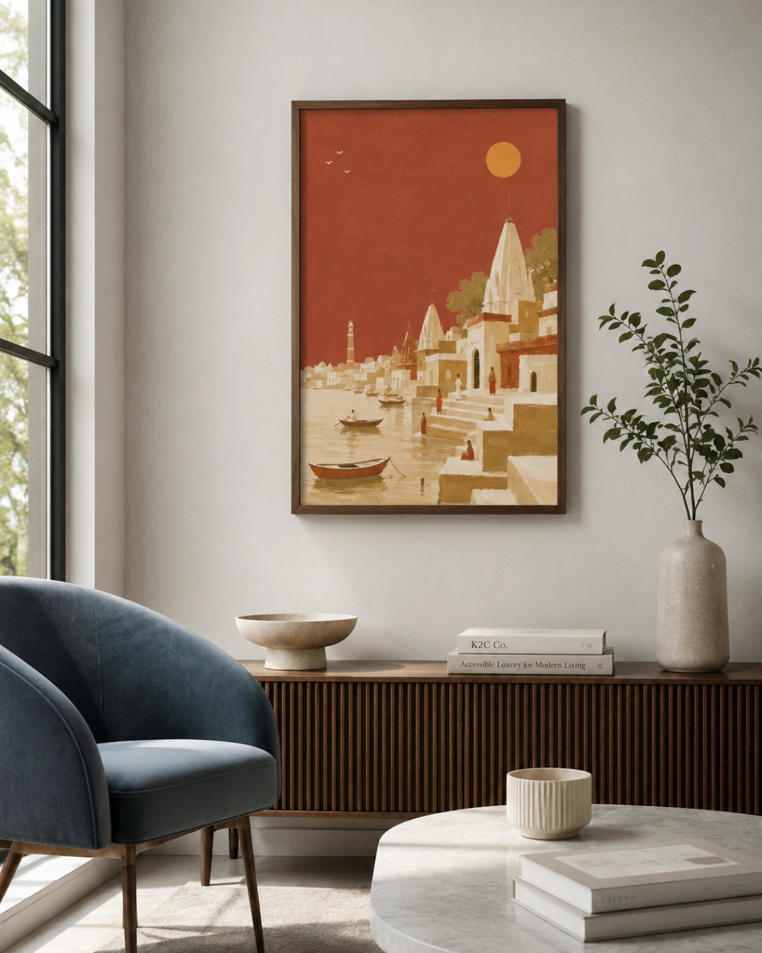 A framed art print depicting a stylized Indian harbor scene with boats and ghats under a warm orange sky, displayed on a light gray wall in a modern living room setting.
