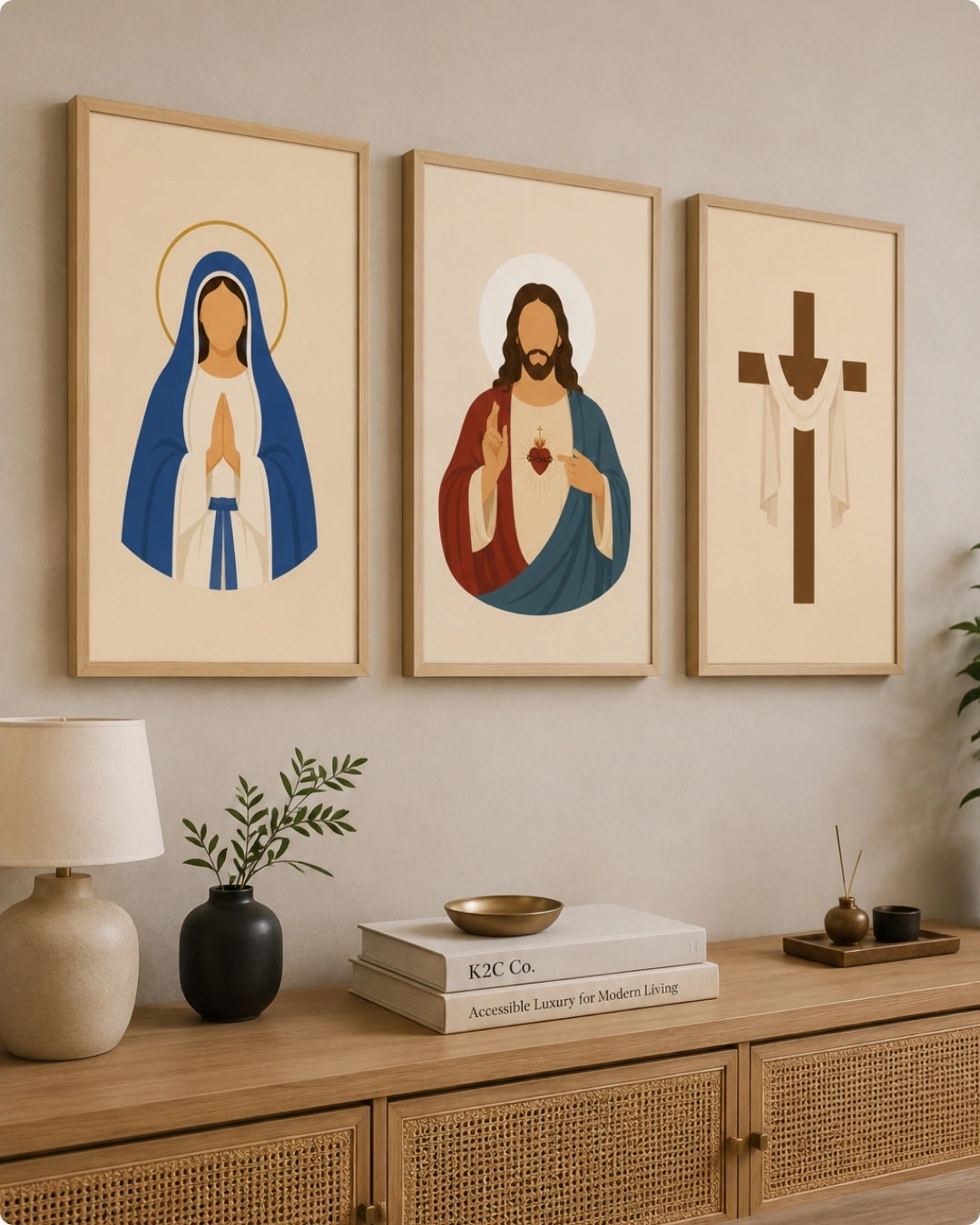 Three framed religious wall art prints hang above a wooden console table in a styled interior. The prints show a robed figure with a halo on the left, a Sacred Heart of Jesus in the center, and a Christian cross on the right, all in a minimalist color palette on warm cream backgrounds.