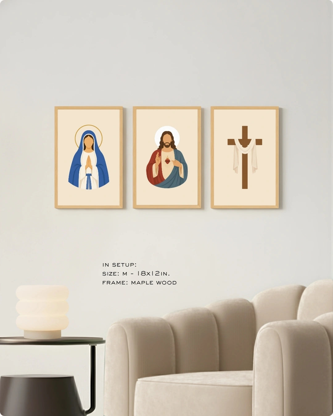 Three framed religious wall art prints hang on a light gray wall above a beige armchair and side table. The prints show a robed figure in blue and white, Jesus with a red heart, and a brown cross, all in light wood frames.