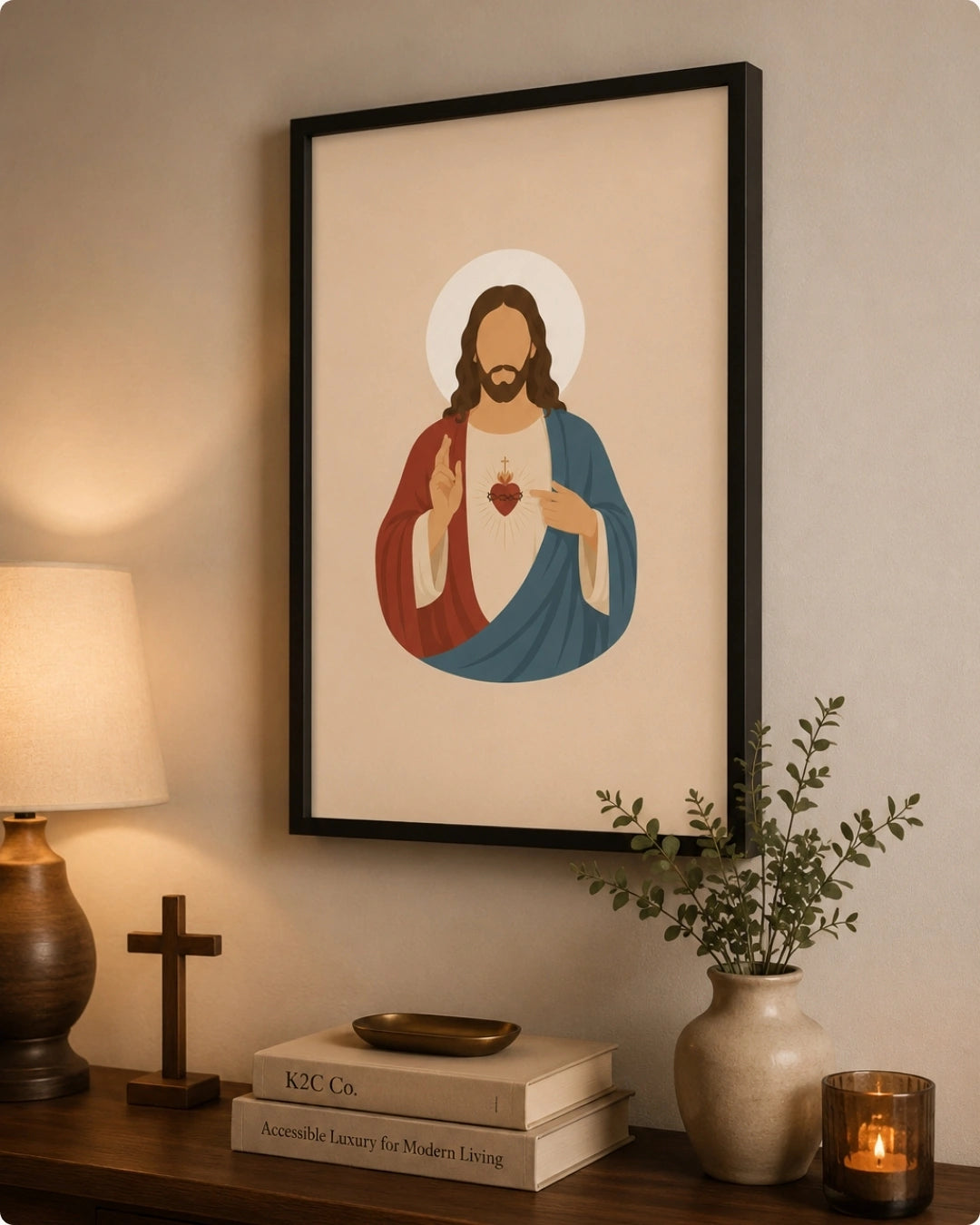 A framed art print of the Sacred Heart of Jesus hangs on a warm beige wall above a wooden console table styled with a lamp, books, a cross, and a vase with greenery.