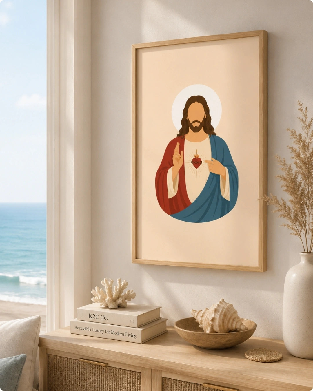 A framed art print of the Sacred Heart of Jesus hangs on a light beige wall in a styled coastal room. The print features a minimalist illustration of Jesus with a red robe, blue cloak, and a glowing heart, set against a warm cream background.