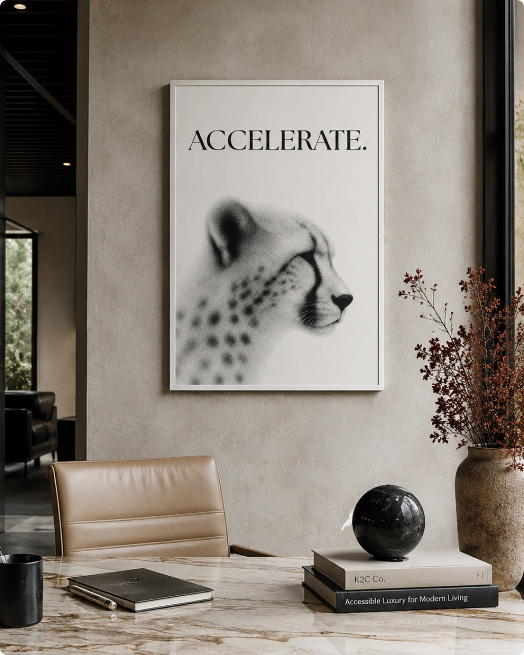 A framed black and white art print featuring a cheetah profile with the word 'ACCELERATE.' above it, displayed on a textured beige wall in a modern interior setting with a desk, chair, and books.