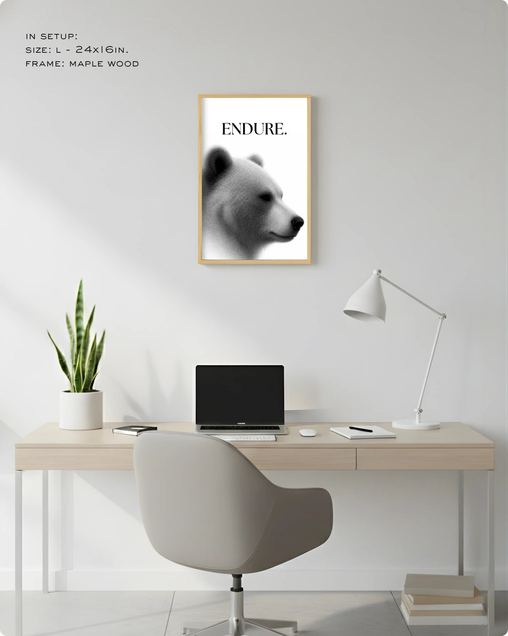 A framed wall art print featuring a black-and-white bear portrait with the word 'ENDURE.' at the top, displayed above a light wood desk in a modern home office setup with a chair, lamp, laptop, and plant.