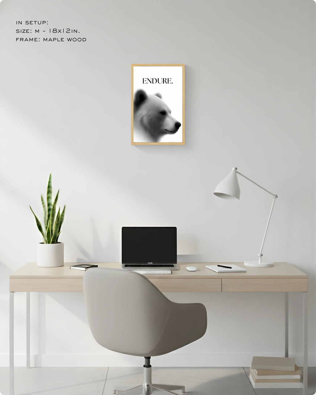 A framed wall art print featuring a black-and-white bear portrait with the word 'ENDURE.' at the top, displayed above a light wood desk in a minimalist home office setup with a chair, lamp, laptop, and plant.